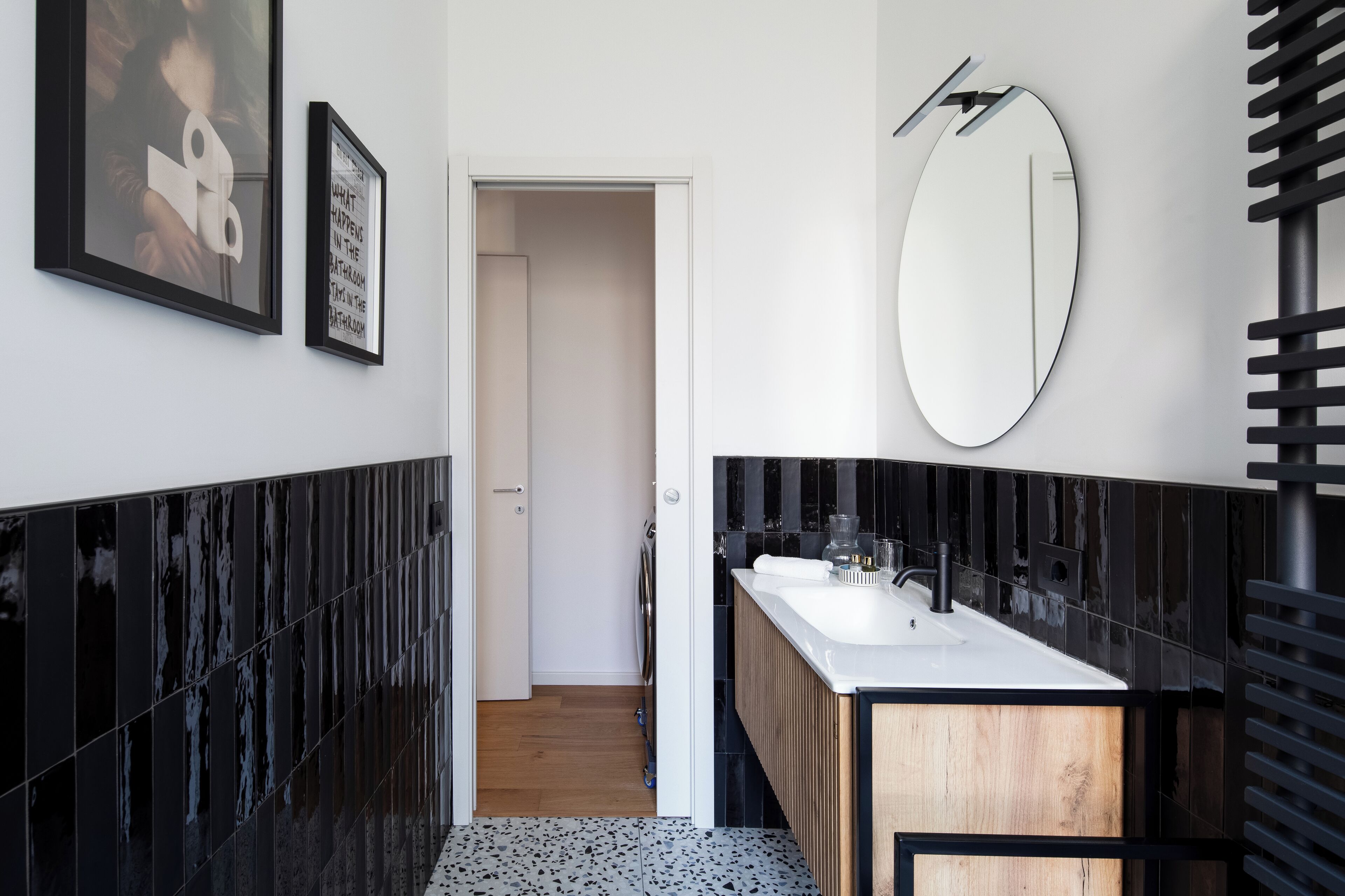 A modern bathroom with black and white tiles, a wooden vanity, a large mirror, and framed artwork on the wall.