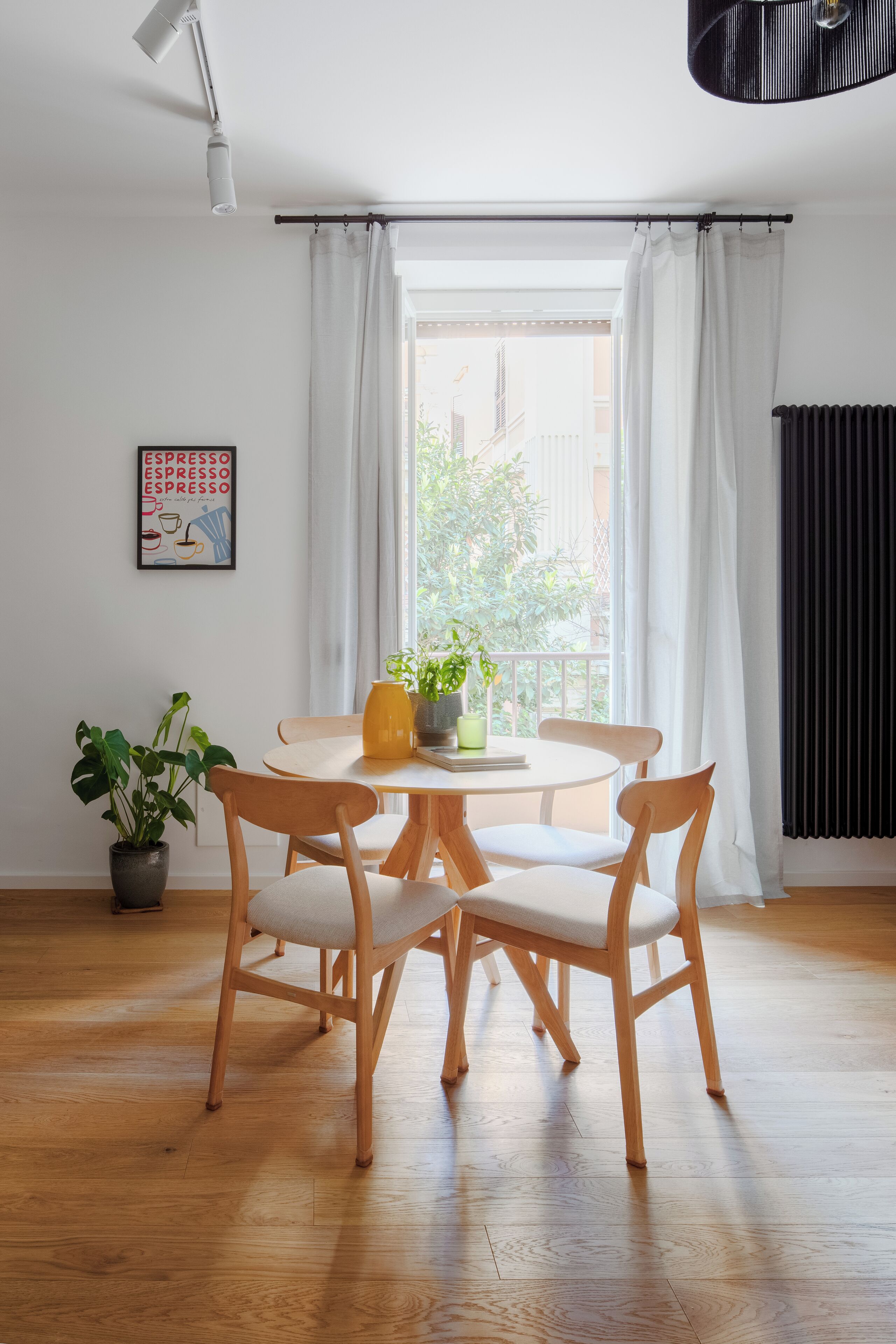 A dining area with a round table, four wooden chairs, a potted plant, and a window with white curtains.