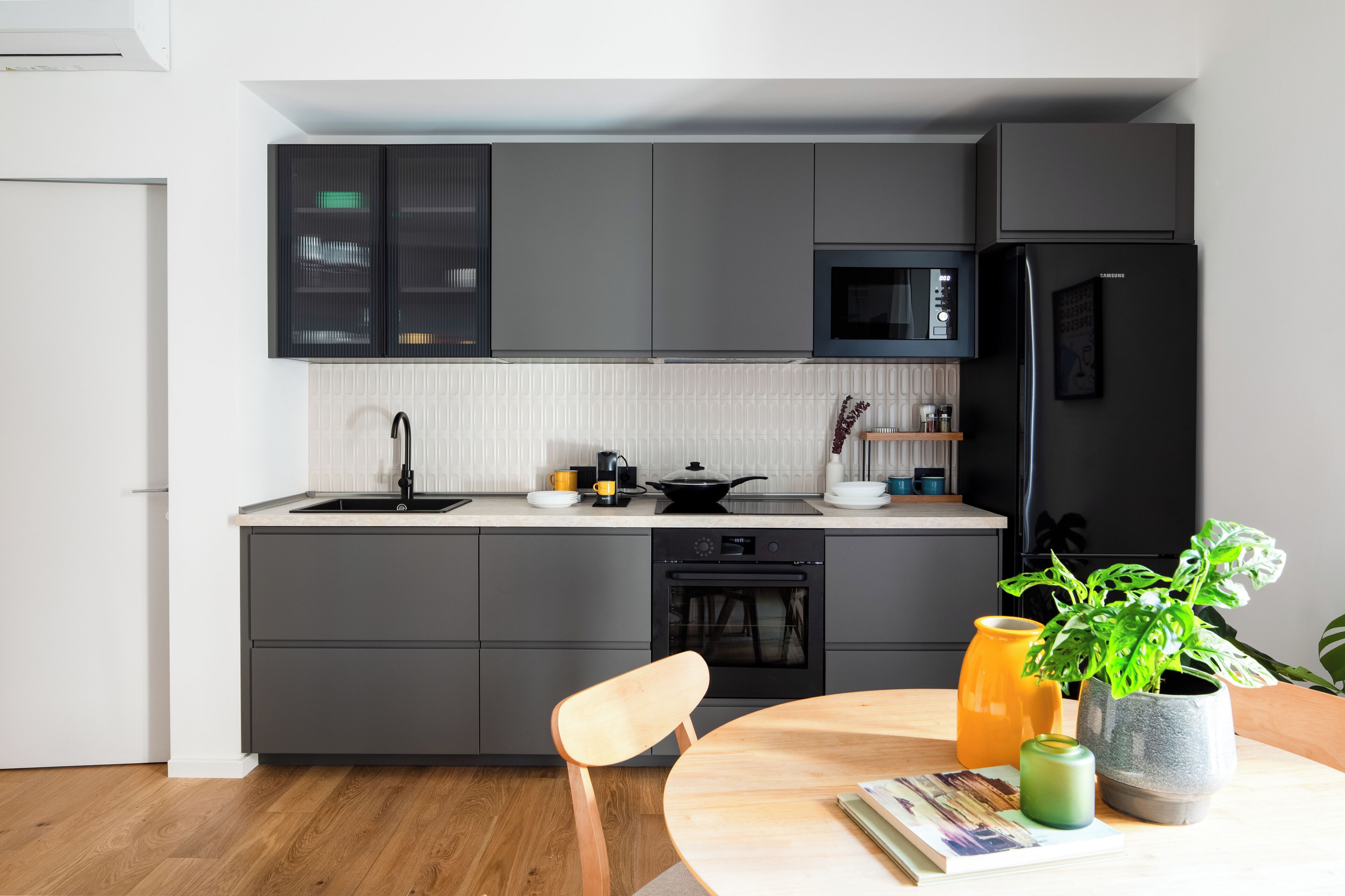A modern kitchen with grey cabinets, a black refrigerator, and a microwave. There is a wooden chair and a table with a plant and a book.
