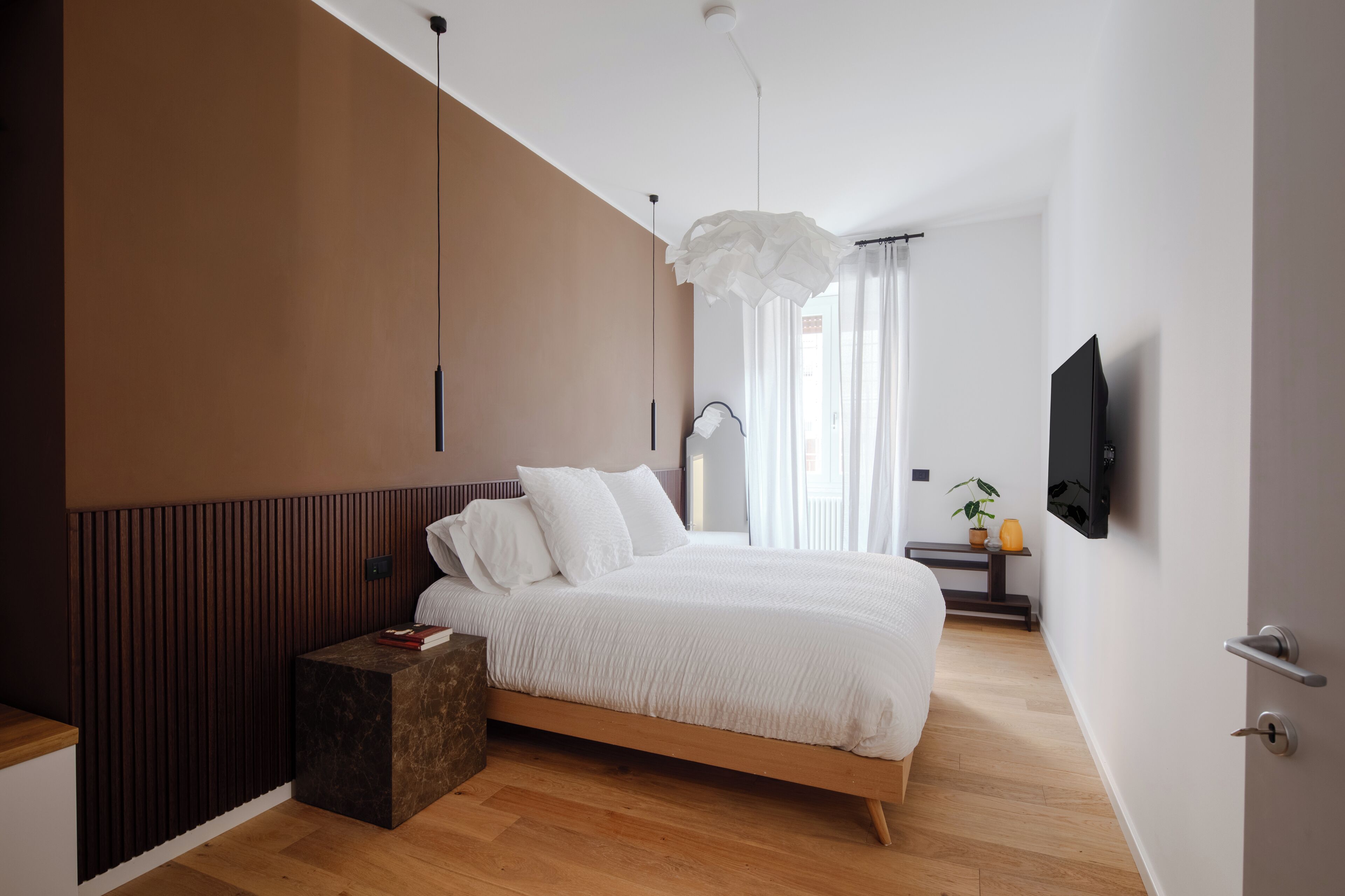A modern bedroom with a wooden bed, a bedside table, a TV mounted on the wall, and a large window with curtains.