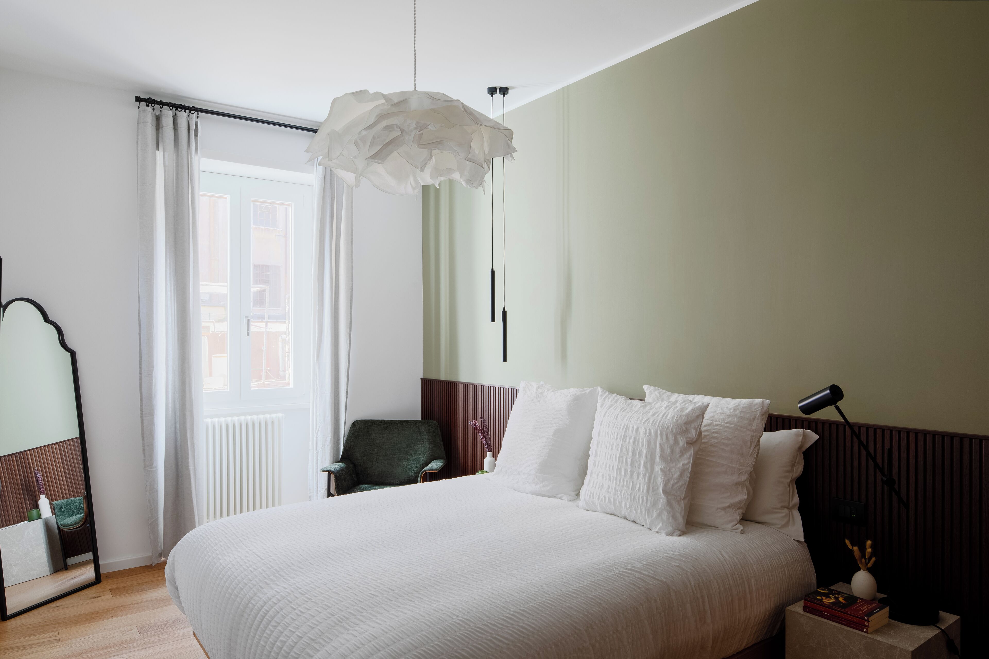 A neatly arranged bedroom with a large bed, a chair, a mirror, and a bedside table with books and a lamp.