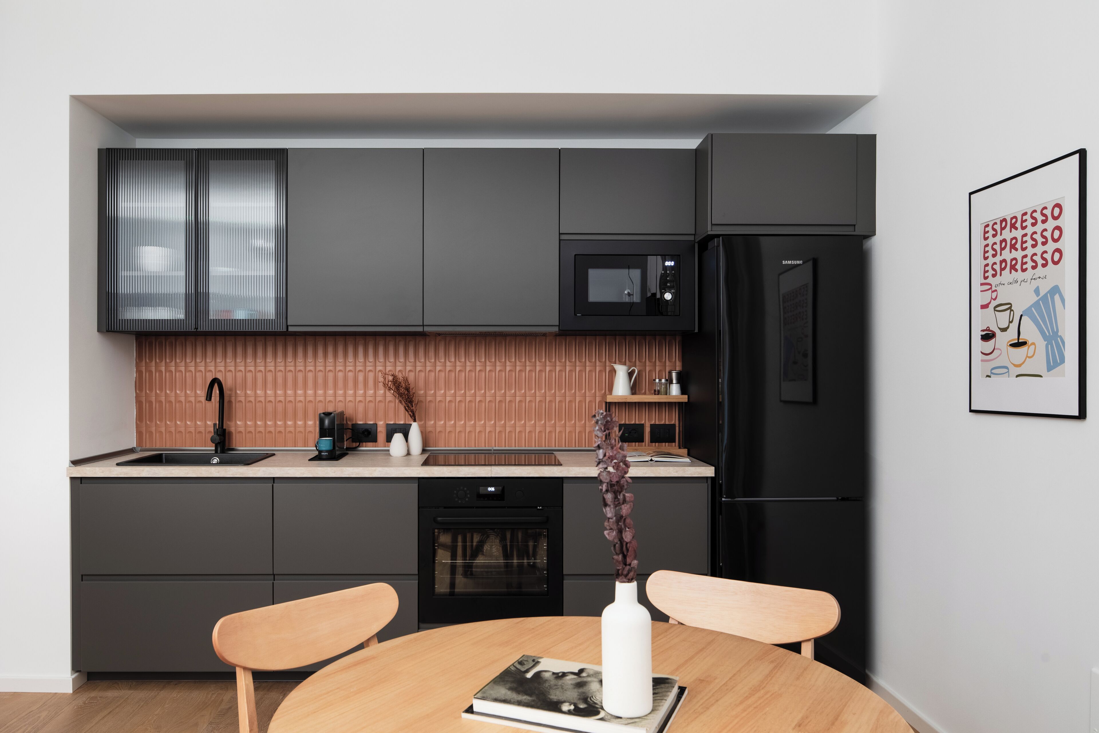 Une cuisine moderne avec des meubles de couleur sombre, un four à micro-ondes et un réfrigérateur. On y trouve une table à manger avec des chaises et un vase rempli de fleurs.