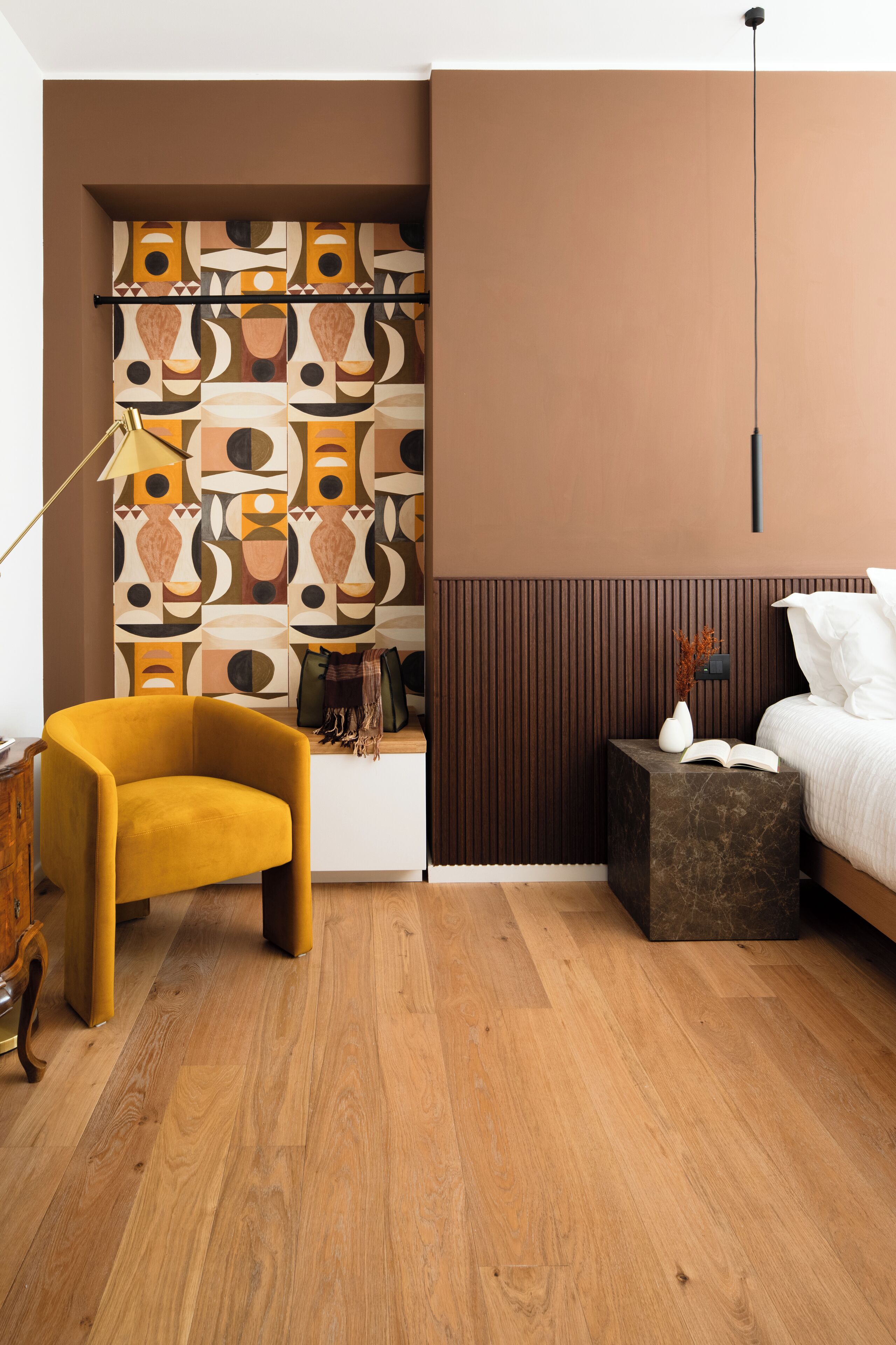 A modern bedroom with a wooden floor, a yellow armchair, a bed with white bedding, a side table with a vase and book, and a wall with geometric artwork.