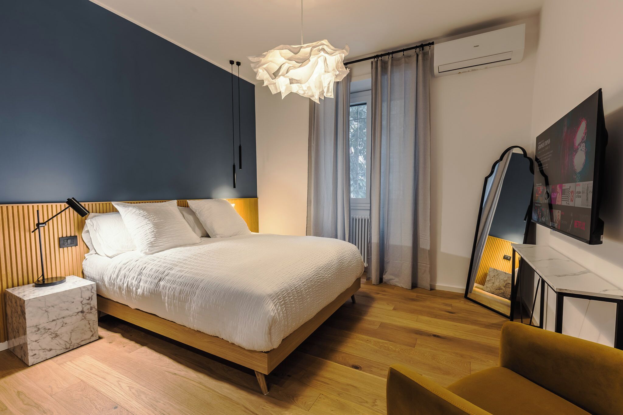 Un dormitorio moderno con una cama grande, una mesita de noche, un espejo, un televisor y una ventana con cortinas.