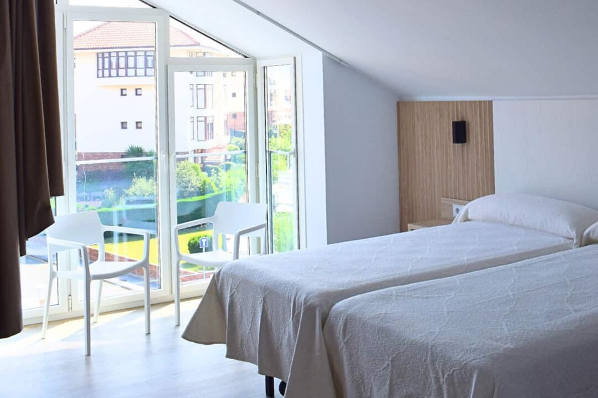 Una habitación de hotel moderna con dos camas, un balcón con una silla y vistas a edificios y zonas verdes.