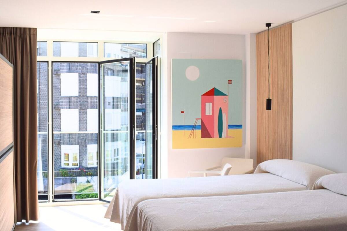 Una habitación de hotel moderna con un gran ventanal que da a un balcón, un cuadro con una escena de playa y una cama con una colcha blanca.