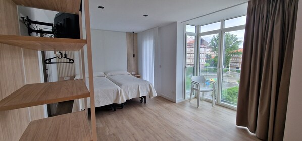 A room with two beds, a window with a view of buildings, a chair, and a shelf with hangers.
