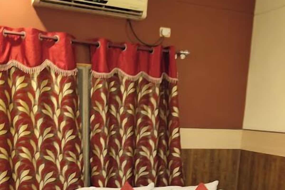 Una habitación de hotel con cama, cortinas rojas, una mesita de noche y un equipo de aire acondicionado.