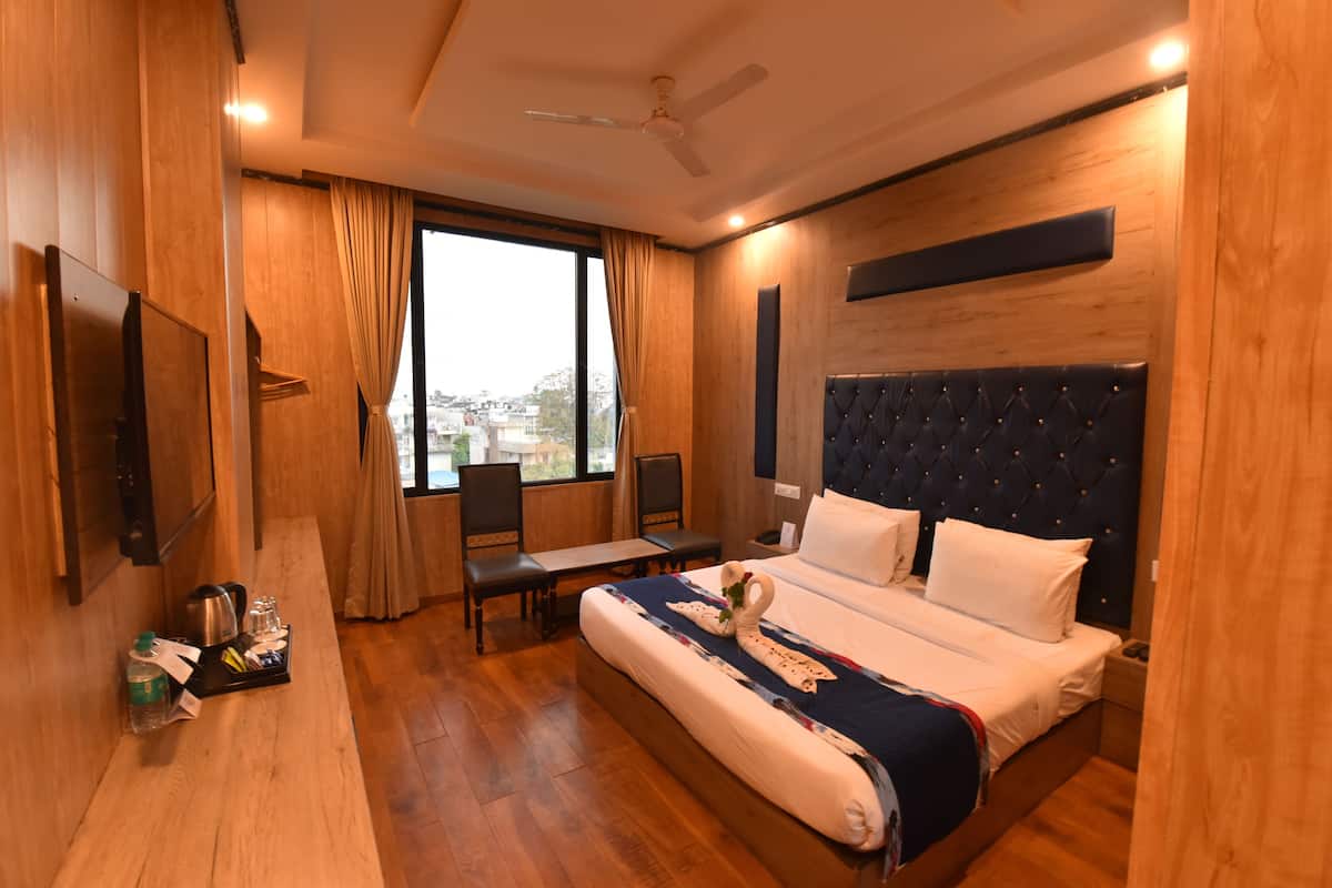 A hotel room with a large bed, a desk with a kettle and glasses, a chair, and a view of the city through the window.