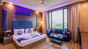 A modern hotel room with a large bed, a blue sofa, a small table, and a large window offering a city view. - Foxoso Fiori Hotel (Jaipur)