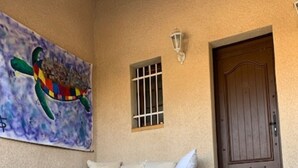 A stone staircase leading to a porch with a gray sofa and white cushions. A wooden door is visible to the right, and a colorful mural hangs on the wall above the sofa. - VILLA POSEÏDON FRONTIGNAN PLAGE (Frontignan)