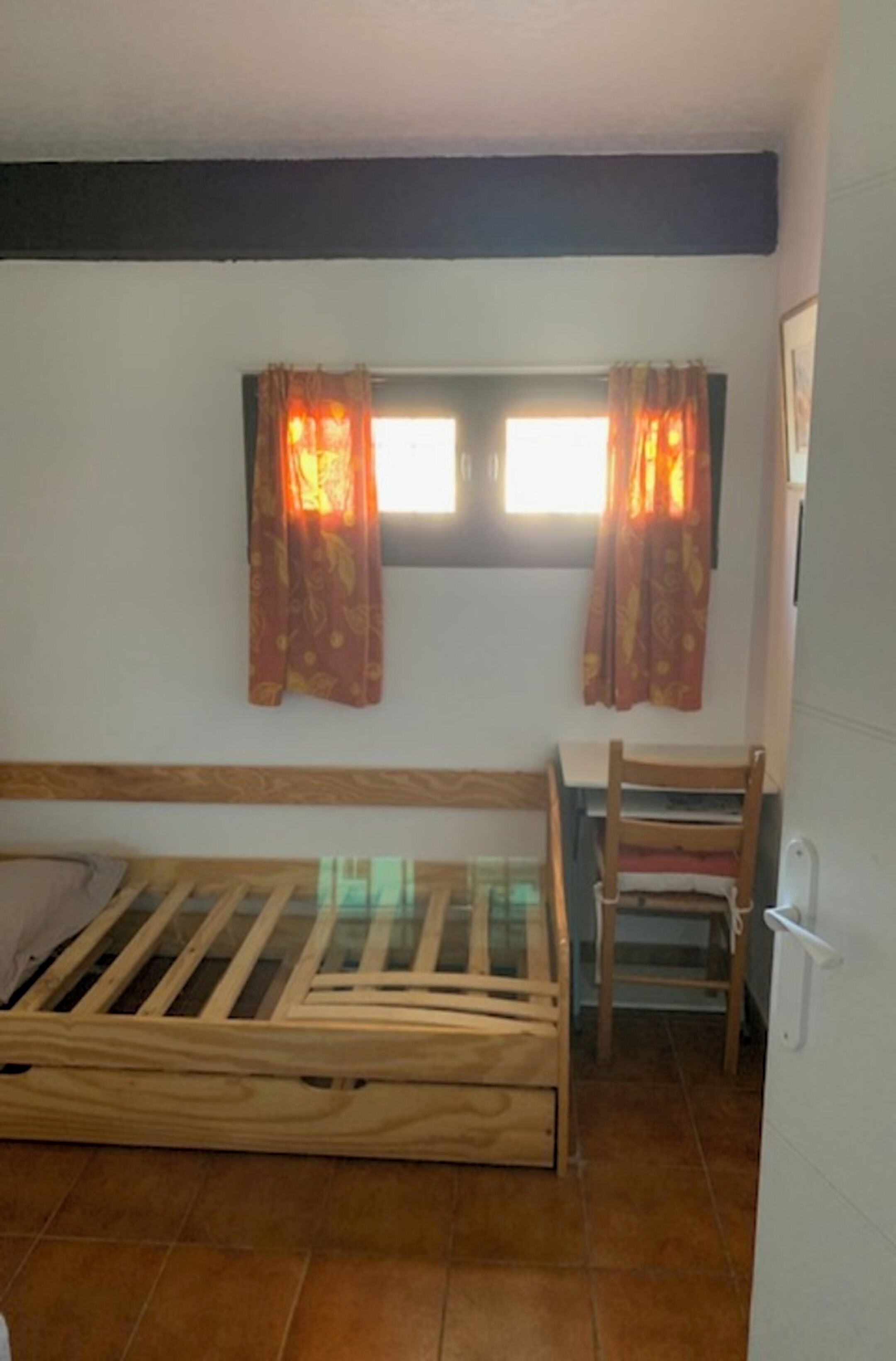 Una habitación pequeña con una cama de madera, una silla y una ventana con cortinas.