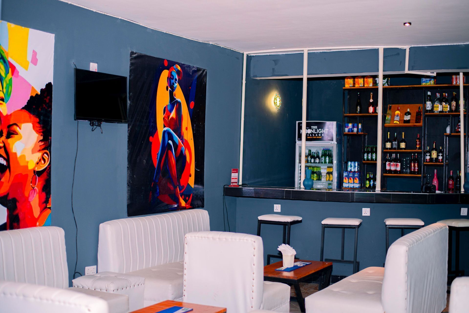 Uma área de bar com assentos estofados em branco, um quadro grande na parede e vista para as prateleiras do bar repletas de garrafas diversas.