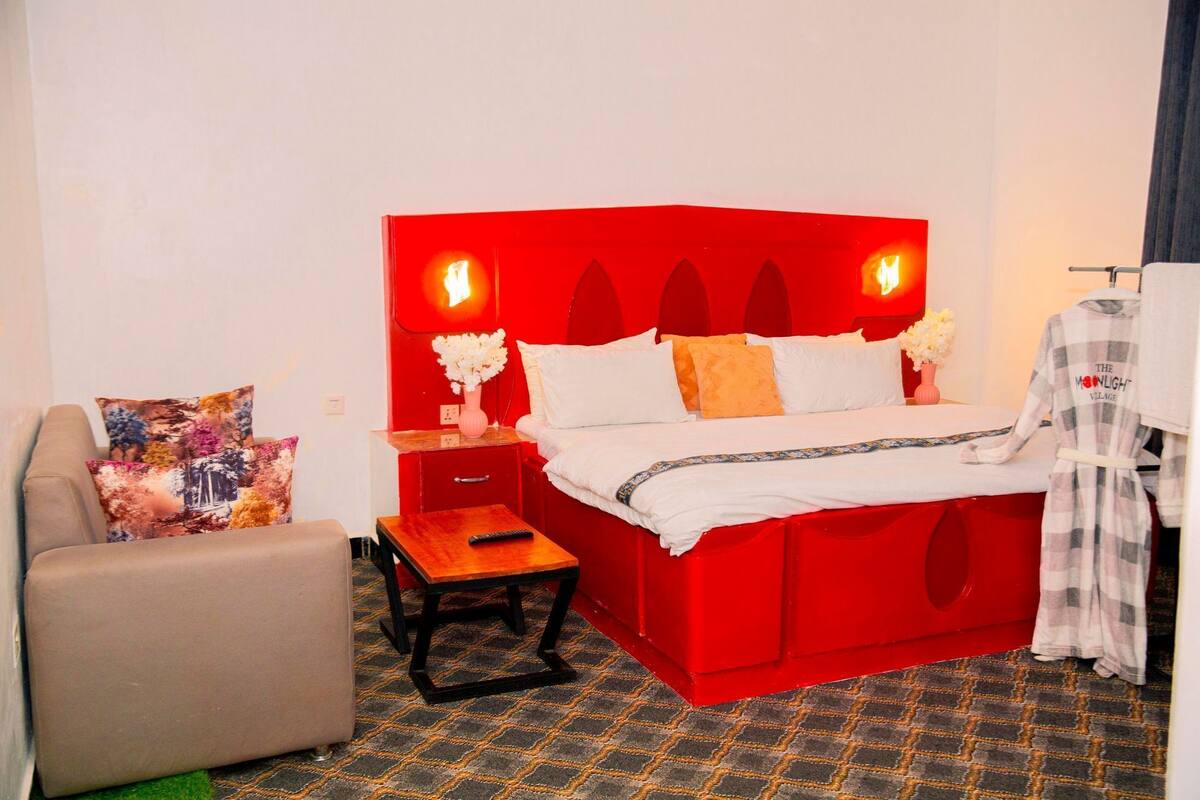 A hotel room with a red headboard, a bed with white and orange bedding, a small wooden table, a chair, and a nightstand with a vase of flowers.
