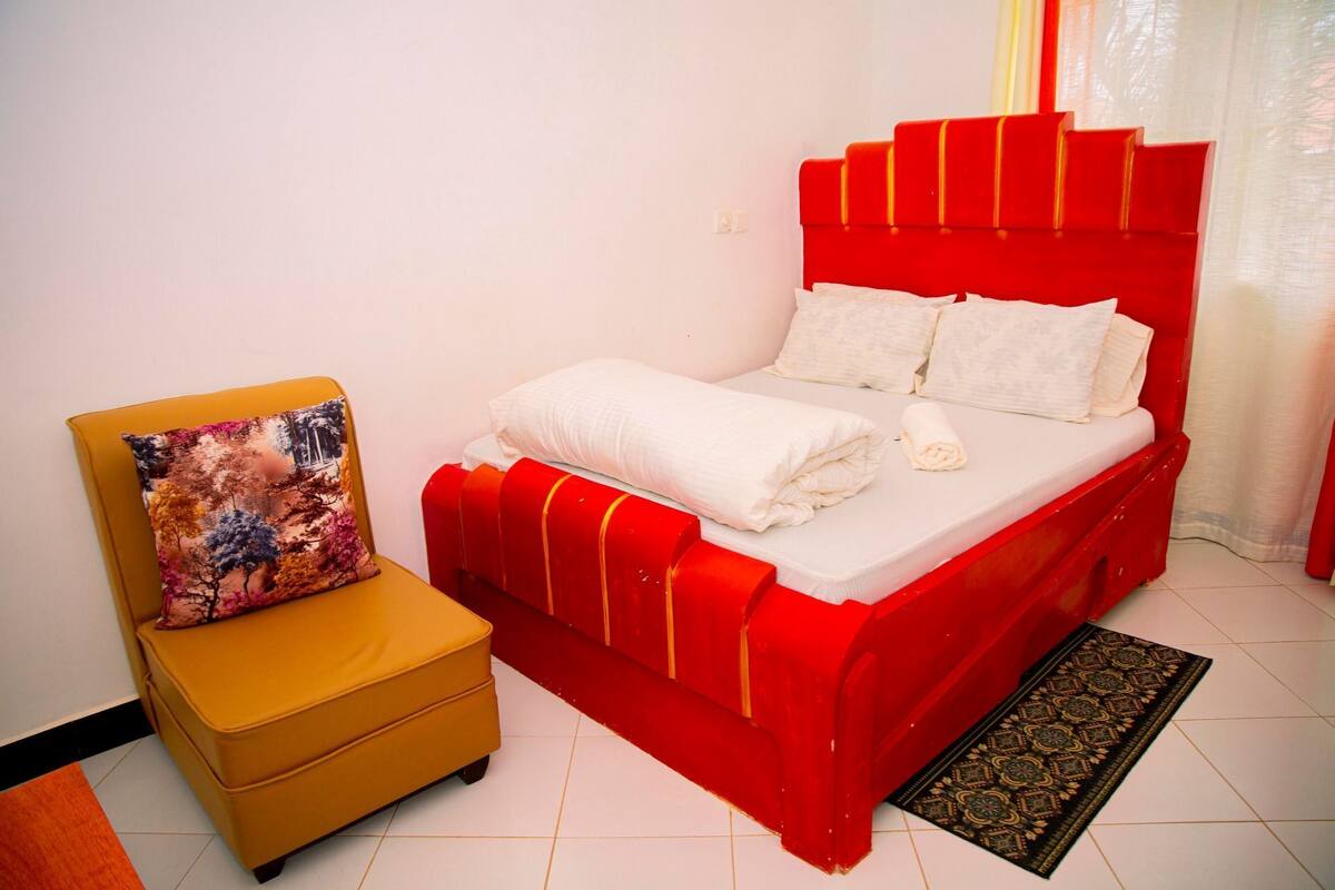 A bed with a red headboard, a yellow armchair, and a patterned pillow.