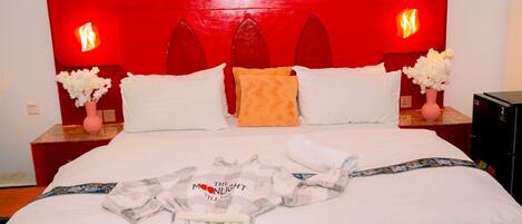 Habitación de hotel con cabecera roja, ropa de cama blanca y una bata sobre la cama. Hay dos mesitas de noche, con jarrones de flores y un dispositivo electrónico negro sobre una de ellas.