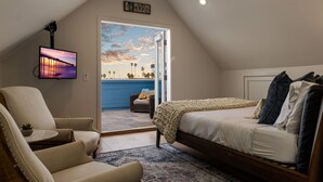 Un dormitorio con cama, una silla, un televisor montado en la pared y vista a la playa a través de una puerta abierta.