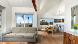Un salón moderno con sofá, mesa de comedor y sillas. El suelo es de madera y hay un ventanal con vista a palmeras y al océano.