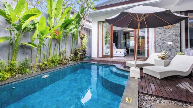 A swimming pool with a wooden deck and lounge chairs, surrounded by lush greenery and a modern house with large windows. - Villa Rama (by JB Villas) (Bali)
