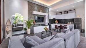 A modern open-plan living area with a grey sofa, a coffee table, and a kitchen with white cabinets and a black backsplash. - Villa Rama (by JB Villas) (Bali)
