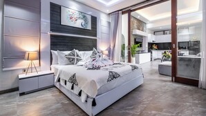 A modern bedroom with a large bed, a nightstand with a lamp, a wall-mounted air conditioner, and a sliding glass door leading to a kitchen area. - Villa Rama (by JB Villas) (Bali)