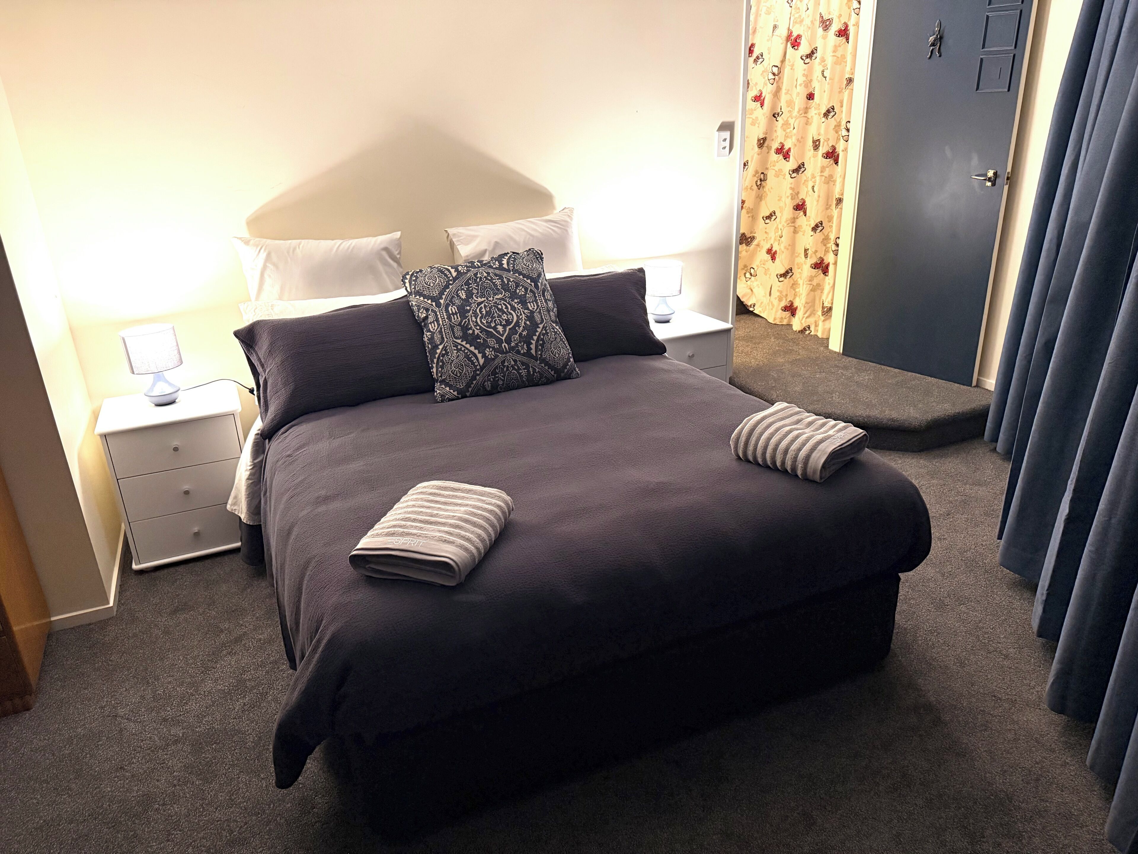 Een opgemaakt bed met een donkere spreidek en kussens, een nachtkastje met een lamp, en een deur met een bloemen gordijn.
