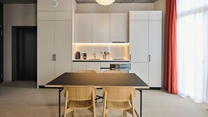 A modern kitchen with white cabinets, a wooden dining table, and chairs. There is a pendant light above the table and a window with curtains on the right. - Ariv Design Apartments (Cham)
