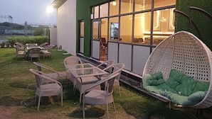 An outdoor seating area with wicker furniture and a hanging chair, a glass table, and a green cushioned sofa. - The Govindam Resort (Vrindavan)