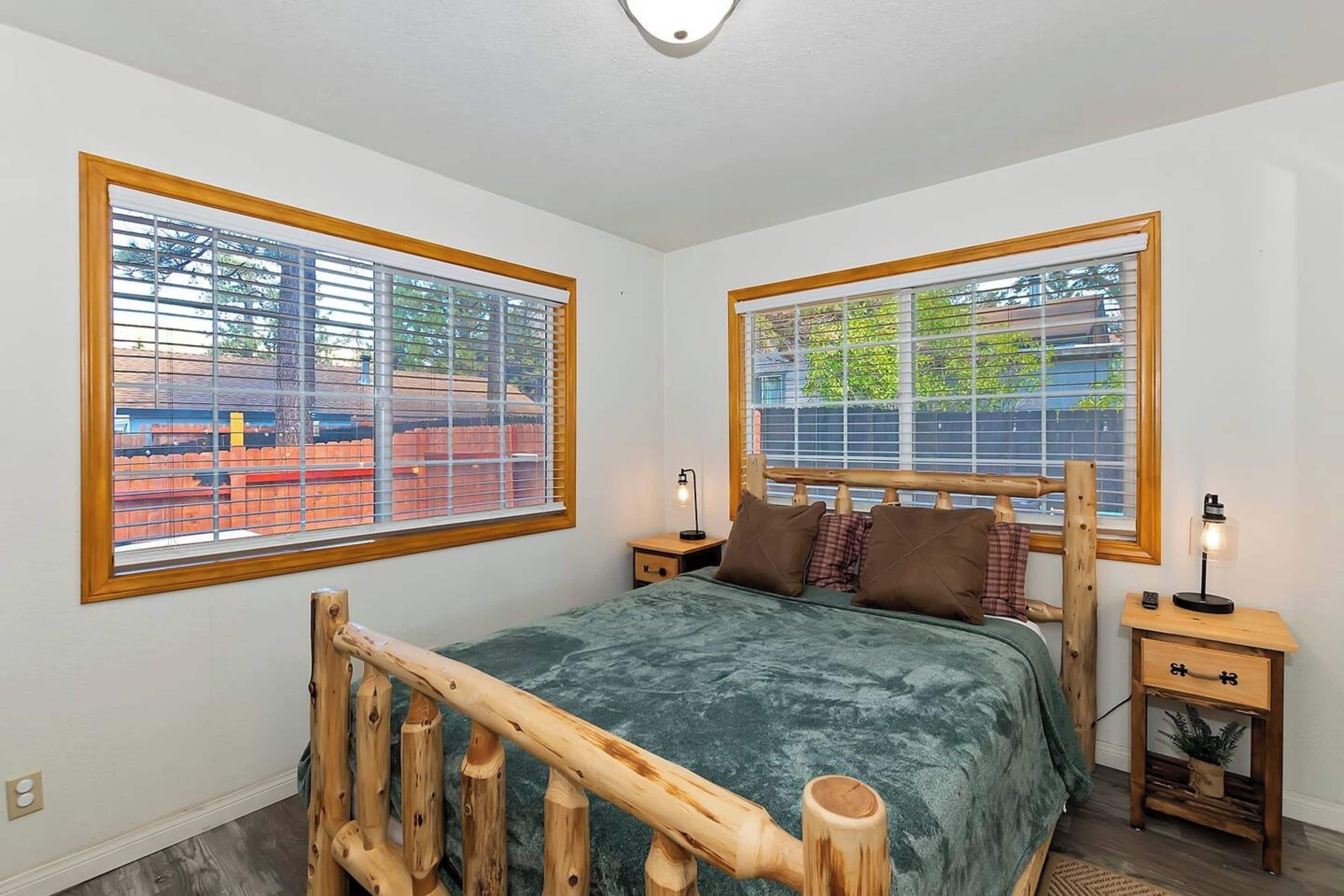 A bedroom with a wooden bed frame, green bedding, two bedside tables with lamps, and large windows with blinds.