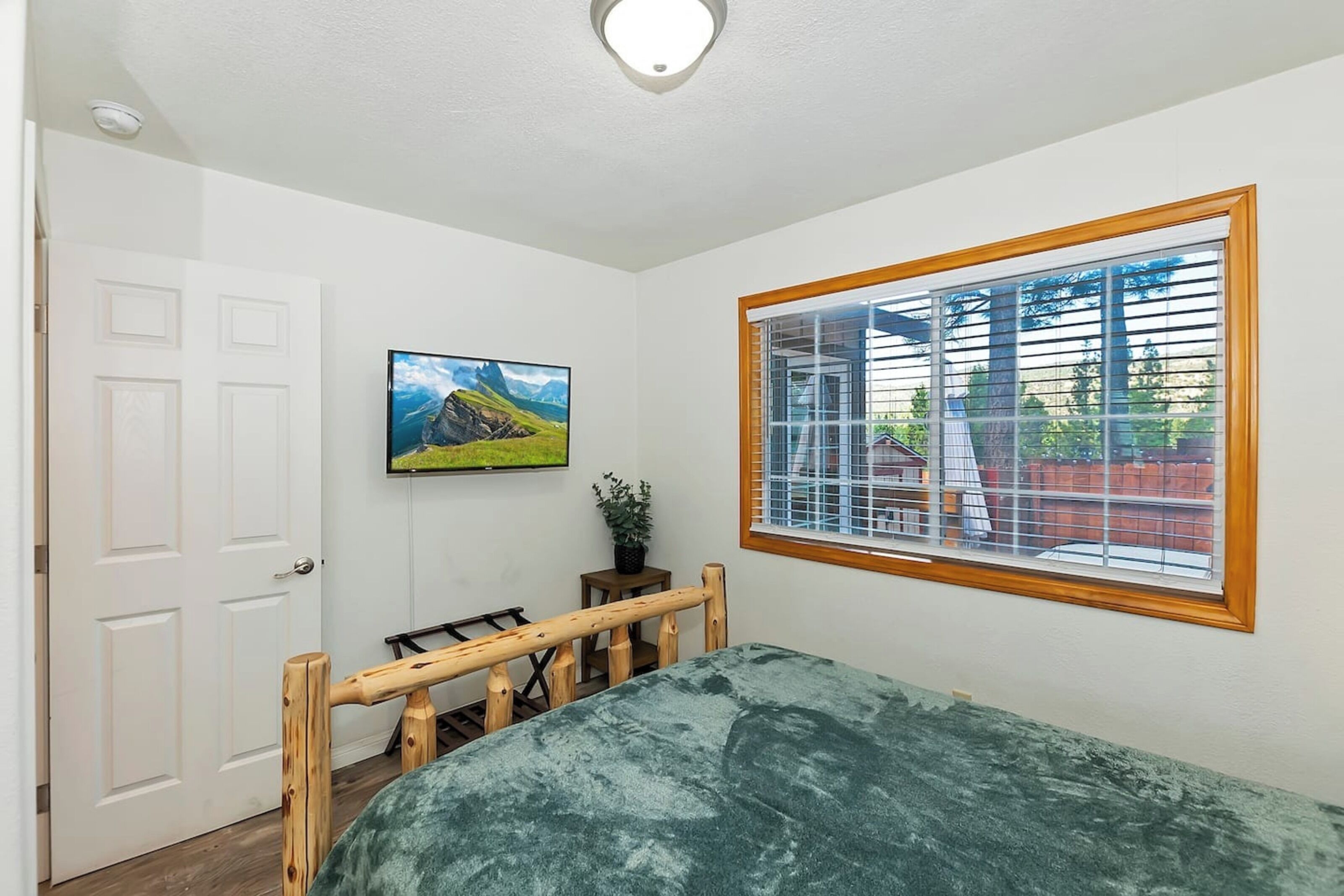 A bedroom with a bed, a window with blinds, a door, and a wall-mounted TV.