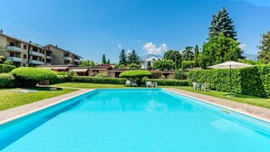 Una piscina rectangular con borde de baldosas, rodeada de setos y árboles bien cuidados, bajo un cielo azul despejado.