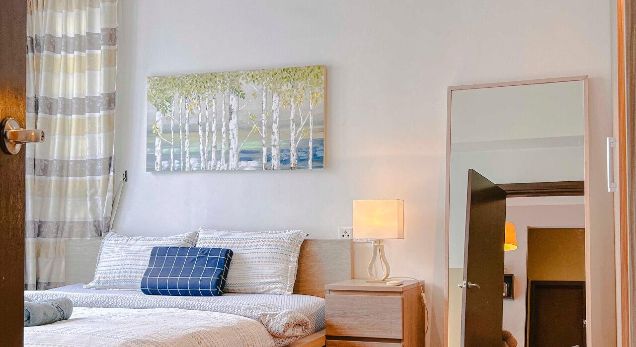 A hotel room with a bed, a nightstand, a lamp, a painting of trees, and a door.