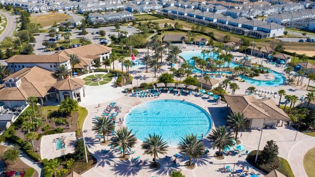 An aerial view of a resort with a large central pool surrounded by palm trees and sun loungers, with residential buildings in the background. - Family-Friendly Home & Pool Near Disney (Davenport)