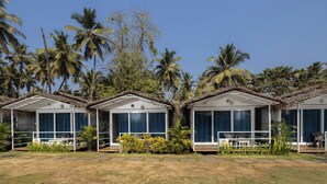 A row of bungalow-style accommodations with large windows, surrounded by palm trees and greenery. - Palolem Casa (Canacona)