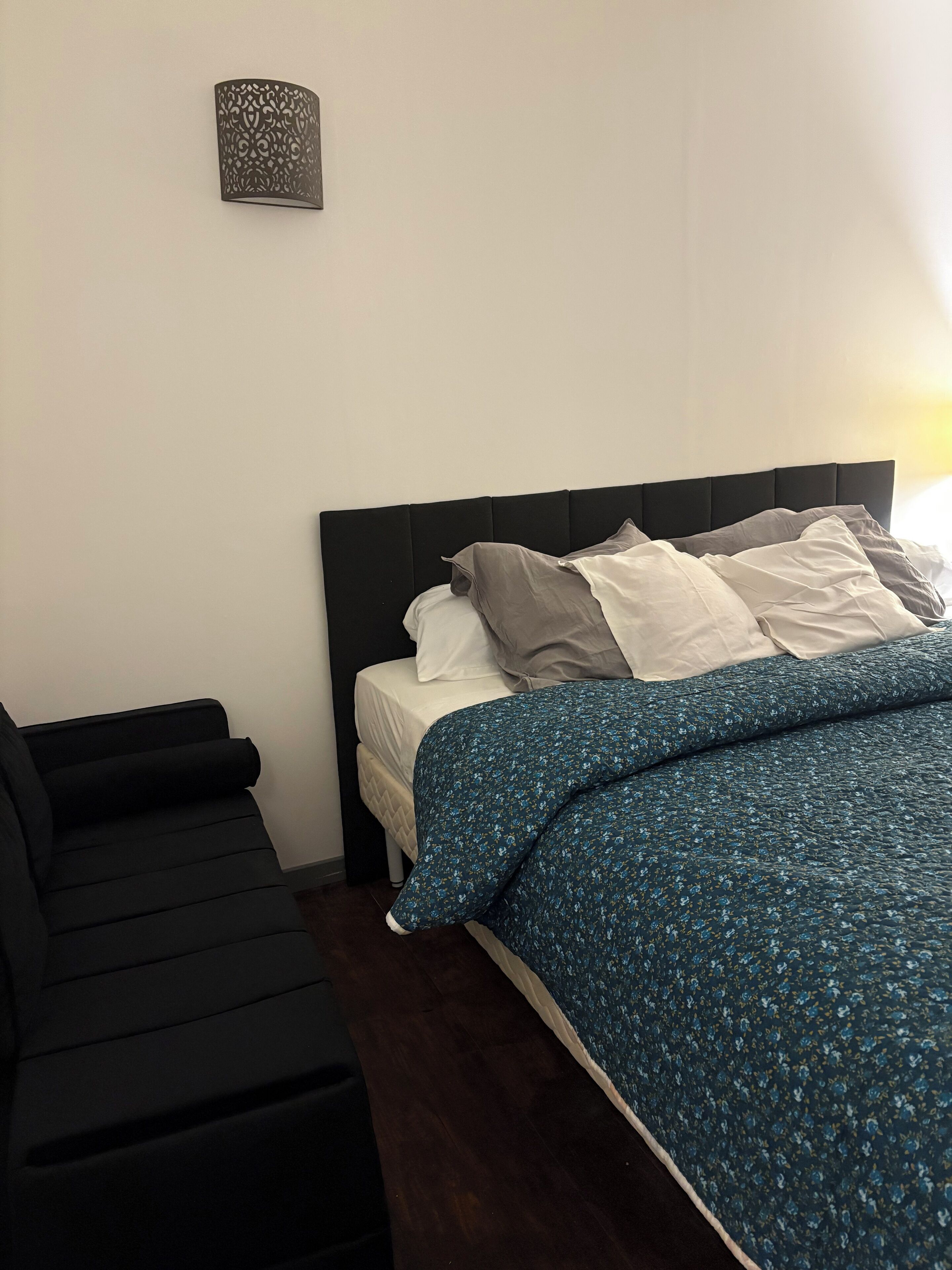A neatly made bed with a blue floral bedspread and white pillows, a black headboard, and a small wall-mounted lamp.