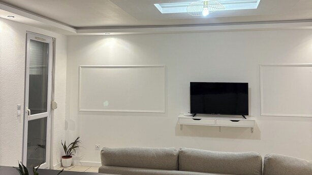 A modern living room with a flat-screen TV, a sofa, a dining table, and a ceiling light fixture.
