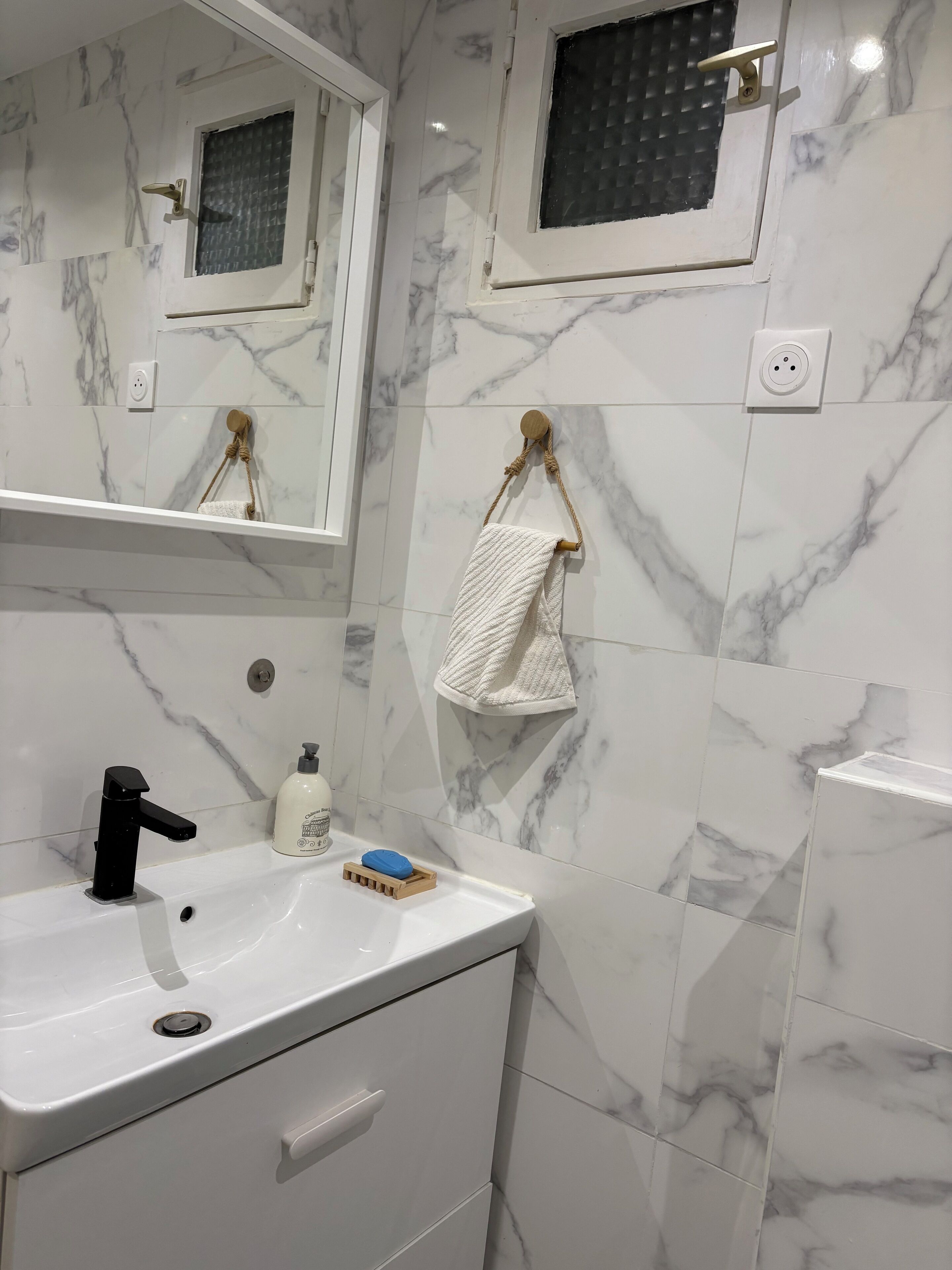 Un baño con lavamanos blanco, espejo, toallero y paredes con diseño de mármol.