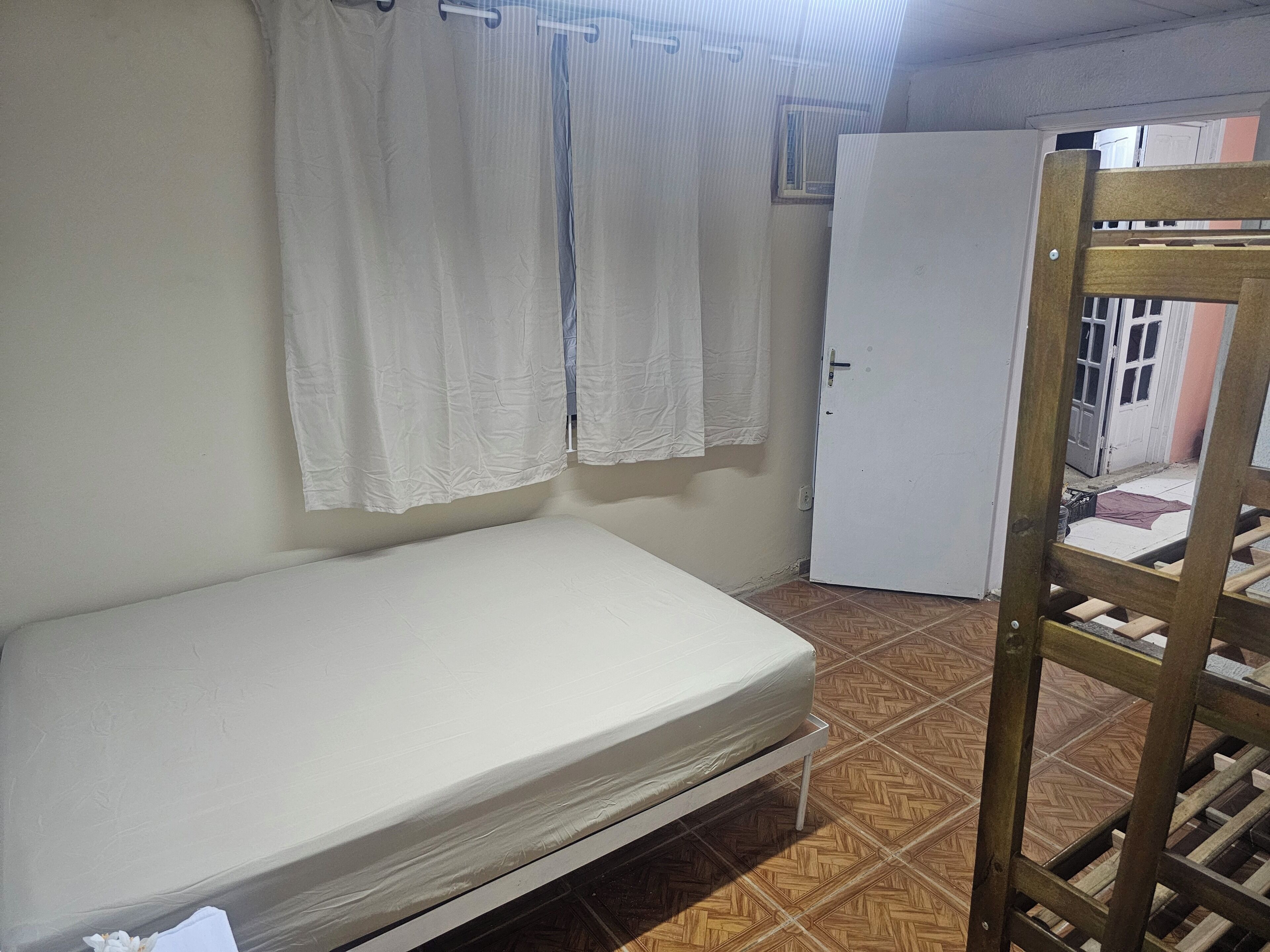 A small, single-bed room with a wooden ladder, white curtains, and a tiled floor.
