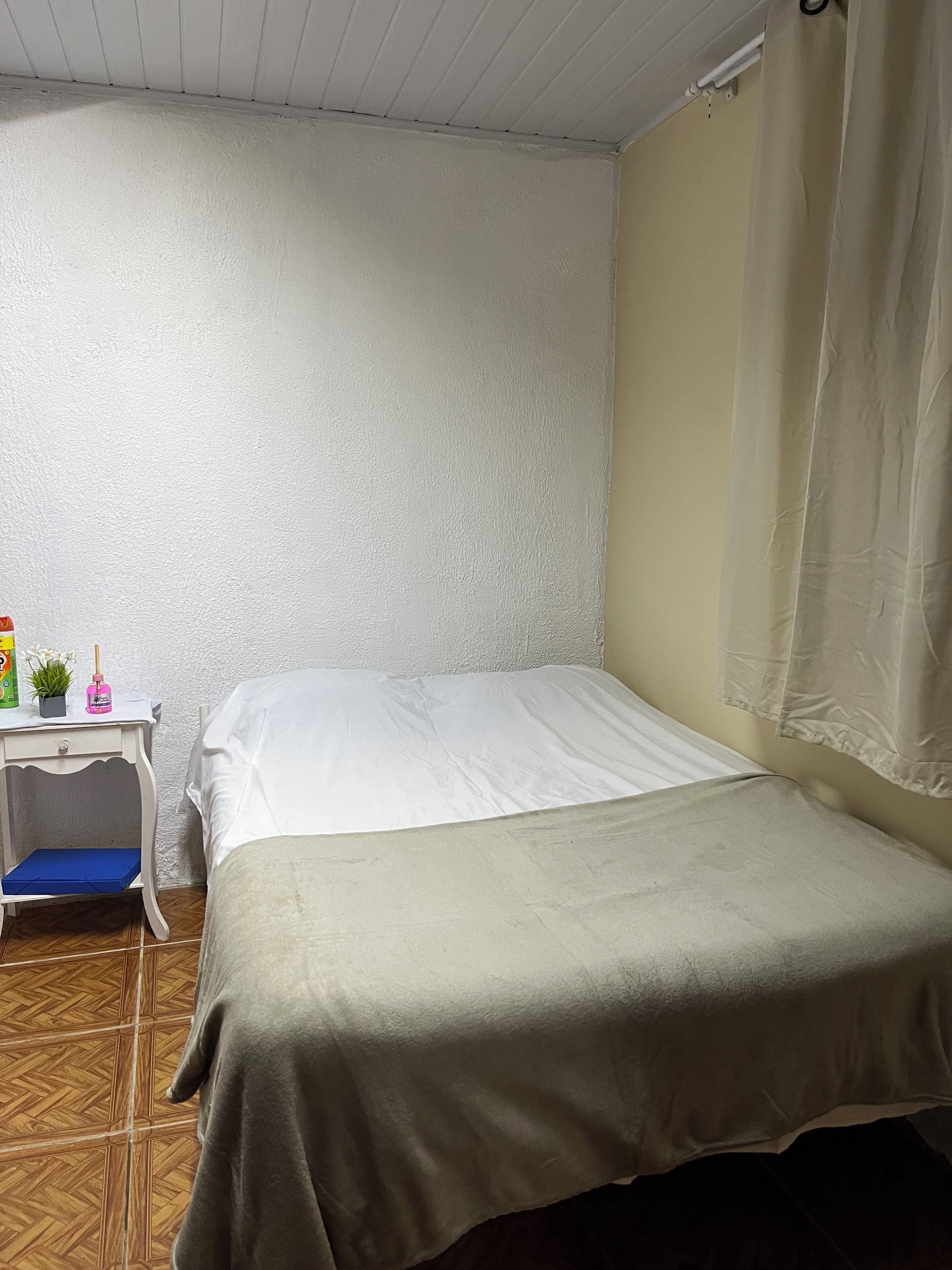 Una habitación pequeña con una cama individual, pared blanca y suelo de madera. Cuenta con una mesita con un cojín azul y algunos objetos encima.