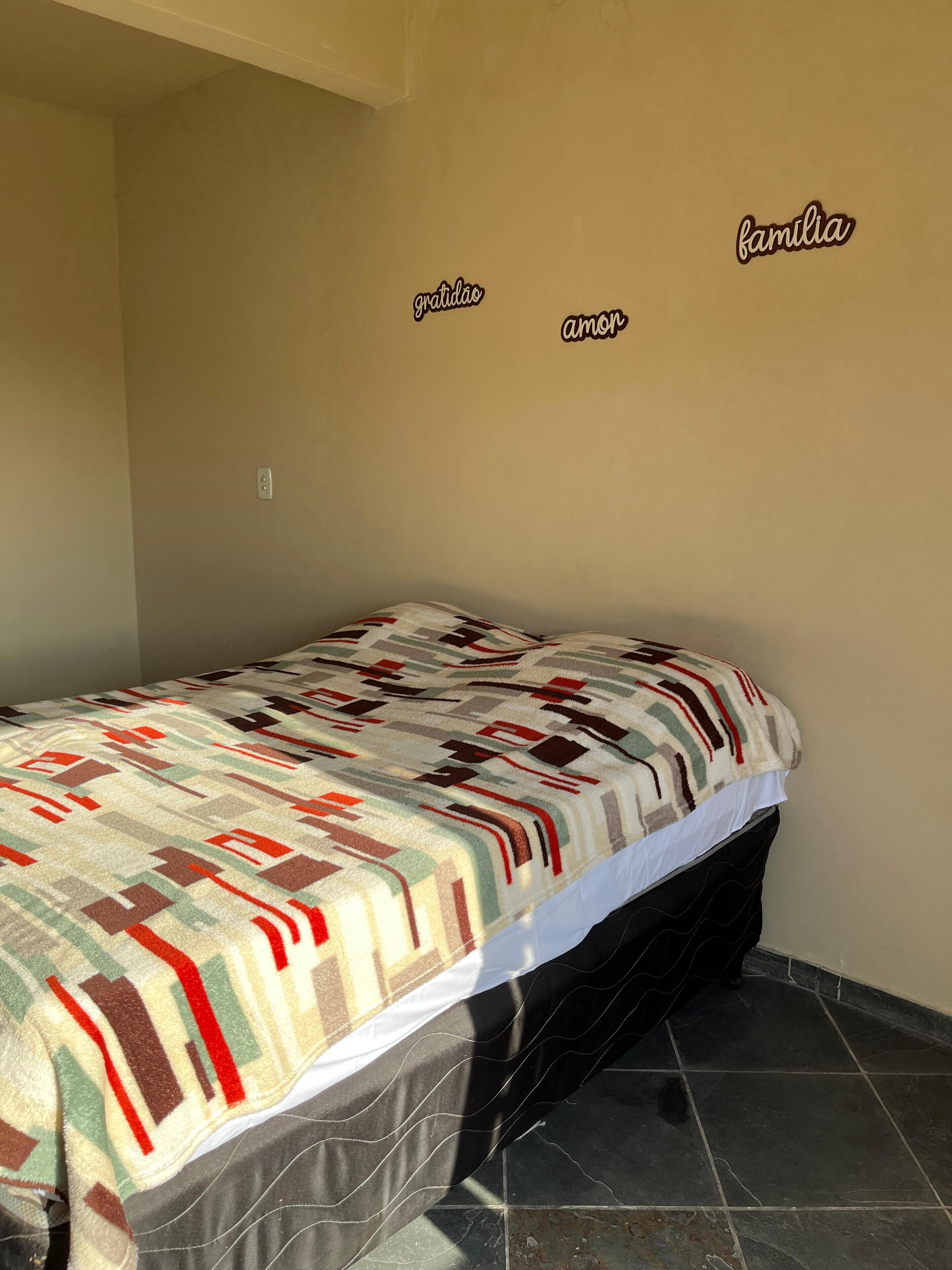Una cama individual con colcha estampada en una habitación con suelo de baldosas y una pared donde se lee 'gratidão amor família'.