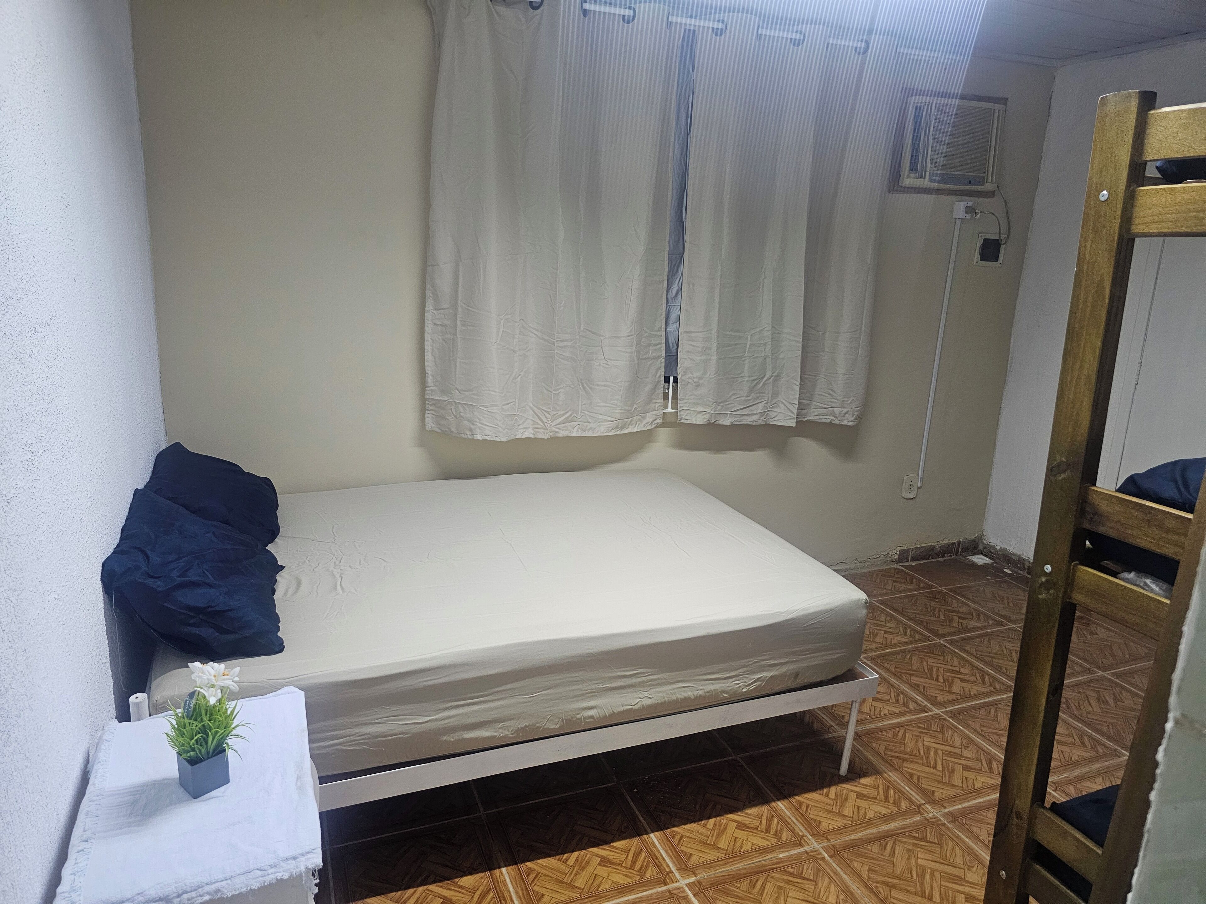 Una habitación pequeña con una cama individual y un catre de madera a la derecha, una planta pequeña en maceta a la izquierda y una ventana con cortinas.