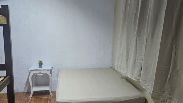 A small, single-bed room with a wooden floor, a white wall, a small white nightstand with a plant, and a window with curtains. - Hostel Sol do Alcaíra  (Arraial do Cabo)