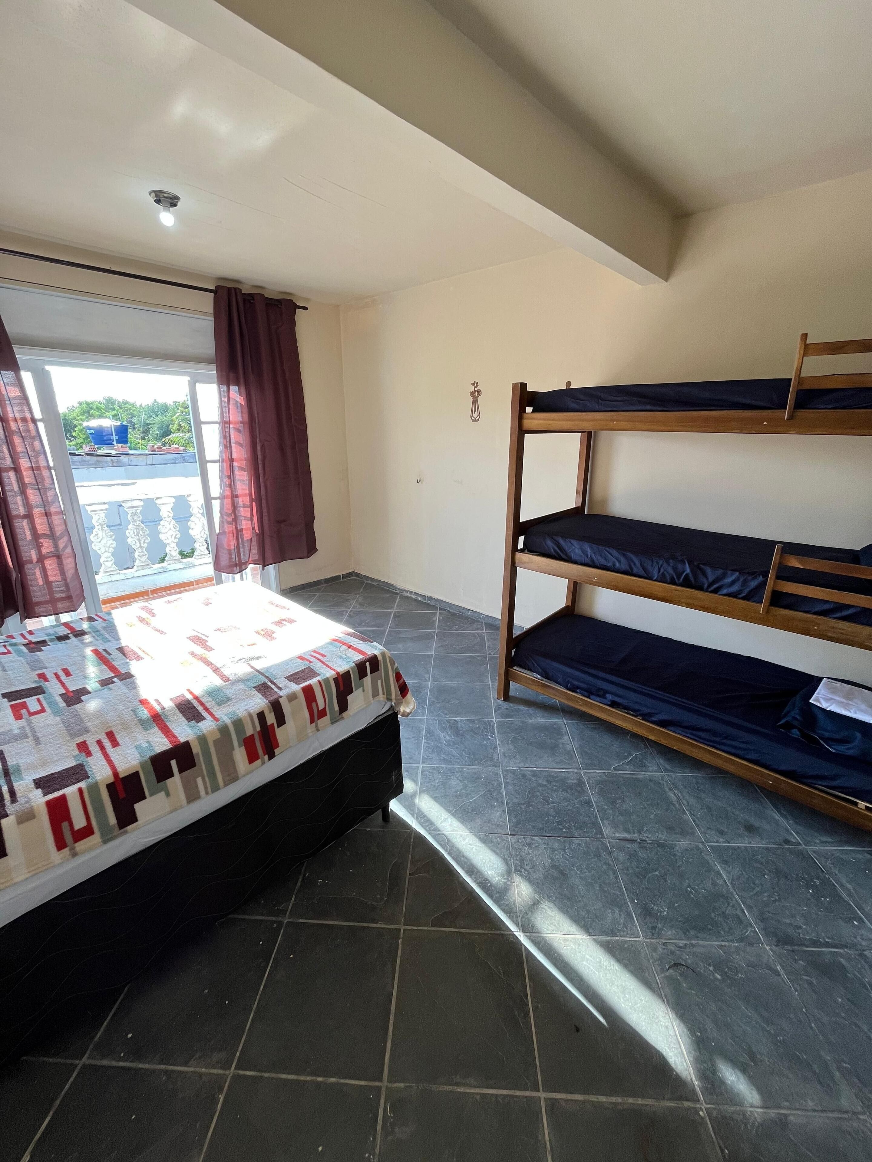 A room with a bed, a bunk bed, a window with curtains, and a view of greenery outside.
