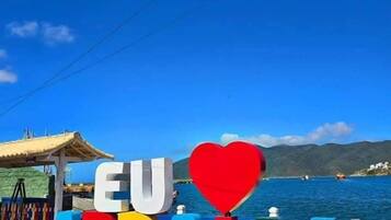 Un letrero colorido con 'EU ARRAIAL' y un símbolo de corazón, con el mar y cielos despejados de fondo.