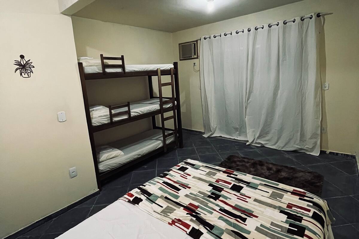 A dormitory room with bunk beds, a single bed, a patterned blanket, a curtain, and a small window.