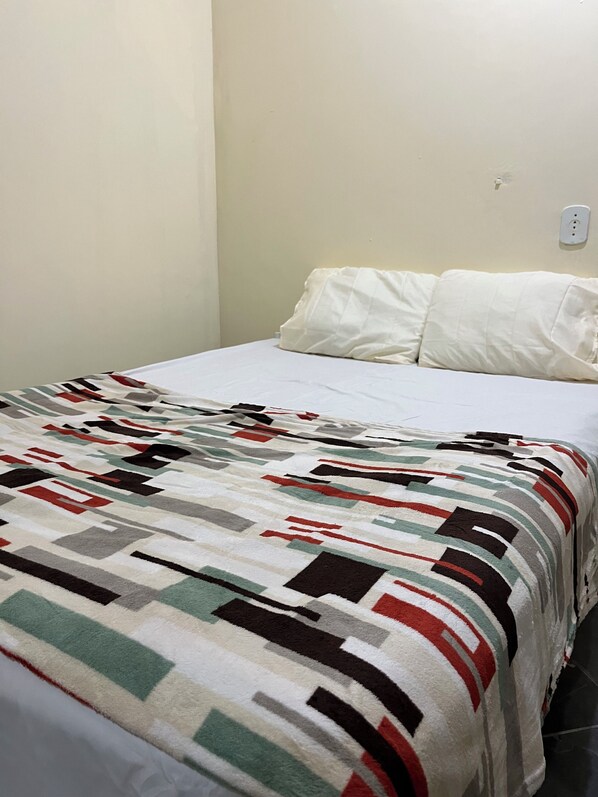 A bed with a patterned blanket, white pillows, and a plain wall with a power outlet.