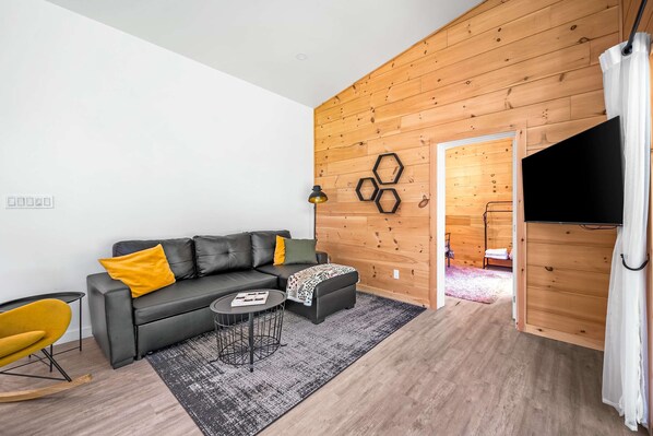 Un salón moderno con un sofá de cuero negro, un sillón amarillo, una mesita redonda y una pared de madera con decoraciones hexagonales.
