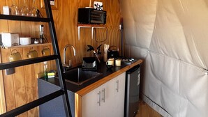 Una cocina compacta con muebles de madera, una encimera negra y un lavamanos pequeño. Hay un horno tostador negro en la encimera, una cafetera y un hervidor. Un tapete a rayas está en el suelo, y una escalera está apoyada contra la pared.