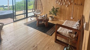 Un interior de madera con un amplio ventanal corredera que da a una terraza, un sillón colgante, un sofá y una cesta de mimbre con plantas secas.