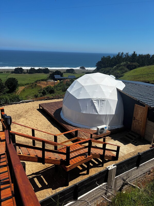 Ein kuppelförmiges Zelt mit Blick aufs Meer und einer hölzernen Terrasse, die darauf führt.